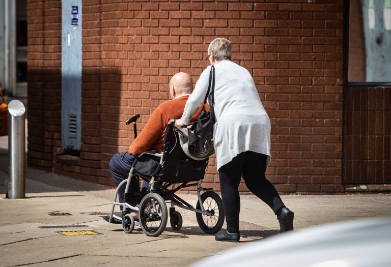 female carer pushing man in wheelchair outside brick building